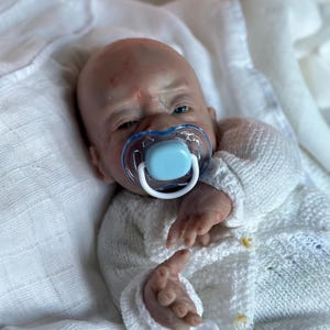 May include: A realistic reborn doll with pale skin, blue eyes, and a bald head, wearing a white knitted cardigan with small buttons. The doll has a light blue and clear plastic pacifier in its mouth and is resting on a soft white fabric.