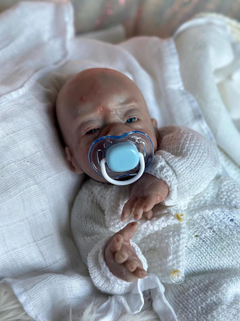 May include: A realistic reborn doll with pale skin, blue eyes, and a bald head, wearing a white knitted cardigan with small buttons. The doll has a light blue and clear plastic pacifier in its mouth and is resting on a soft white fabric.