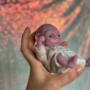 May include: A miniature baby doll with a purple complexion, dressed in a white nappy and a cream knitted jumper. The doll has detailed facial features and is approximately 15 cm long.