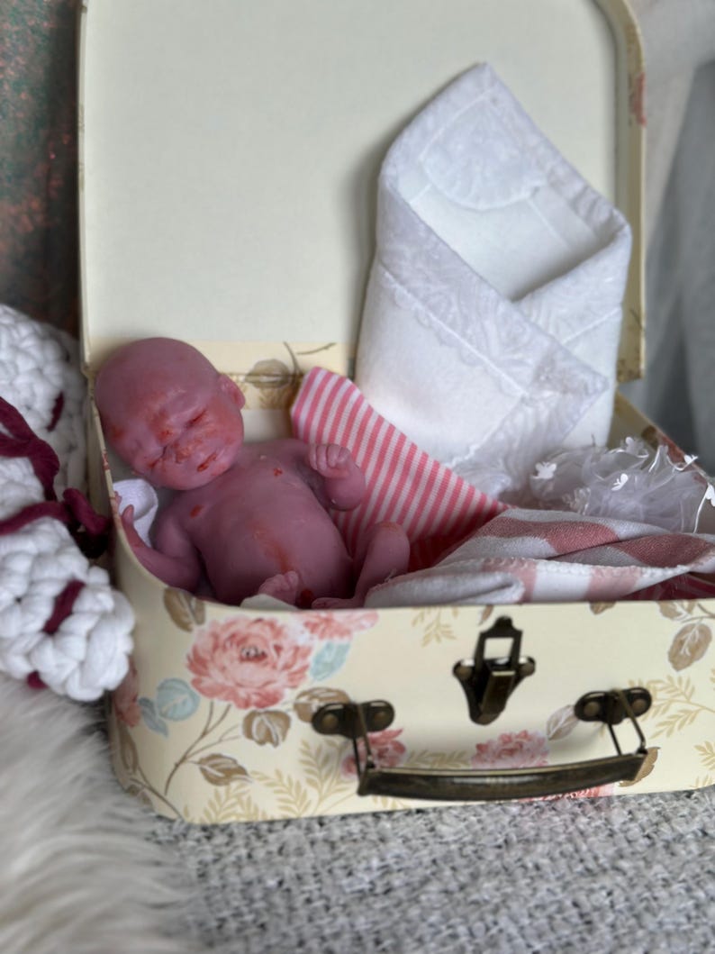 K&ouml;nnte beinhalten: Eine kleine, rosafarbene Silikonpuppe in einem cremefarbenen Miniaturkoffer mit Blumenmuster. Der Koffer enth&auml;lt ein wei&szlig;es, mit Spitze verziertes Tuch und einen rosa-wei&szlig; gestreiften Stoff sowie einen Metallverschluss.