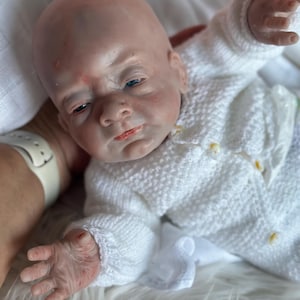 May include: A realistic reborn baby doll with pale skin, blue eyes, and subtle red mottling on the face. The doll has a bald head and is wearing a white, long-sleeved, textured knit cardigan sweater with small buttons down the front.