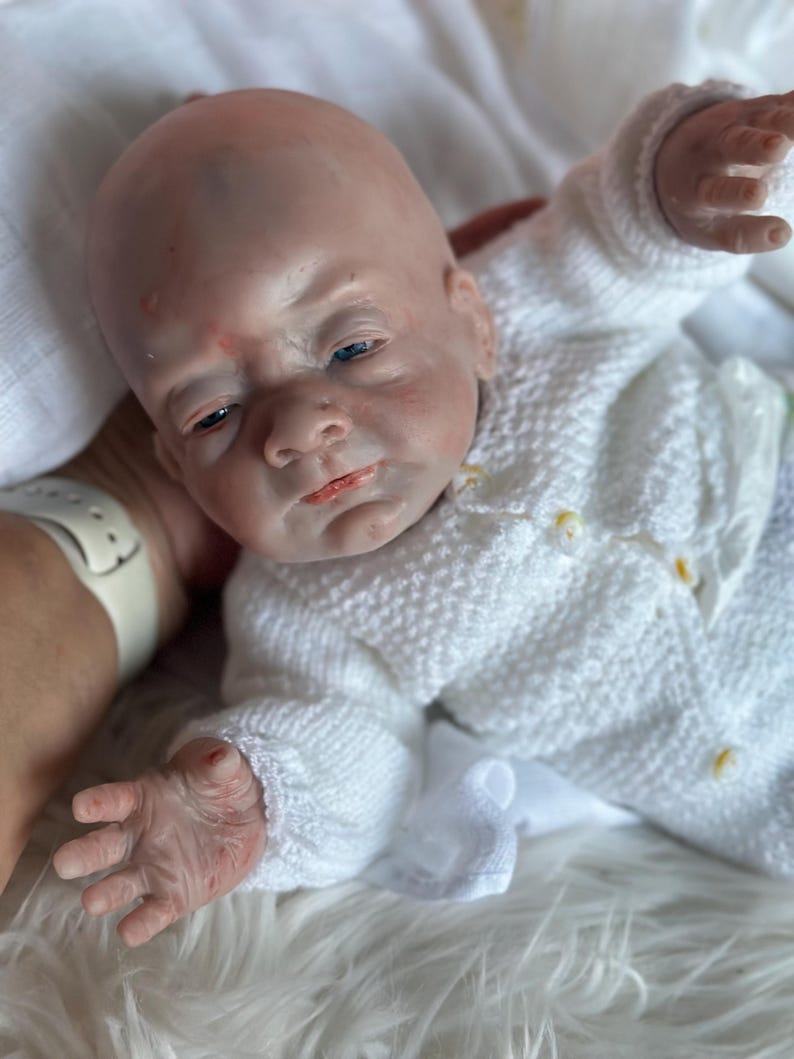 May include: A realistic reborn baby doll with pale skin, blue eyes, and subtle red mottling on the face. The doll has a bald head and is wearing a white, long-sleeved, textured knit cardigan sweater with small buttons down the front.
