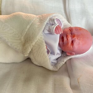K&ouml;nnte beinhalten: Eine realistische Silikonpuppe mit rosafarbenem Teint, geschlossenen Augen und kleinem Mund, eingewickelt in ein wei&szlig;es lang&auml;rmeliges Kleidungsst&uuml;ck und eine wei&szlig;e Wickeldecke mit Spitzenbesatz, mit einem glatten, haarlosen Kopf und sichtbaren Fingern.