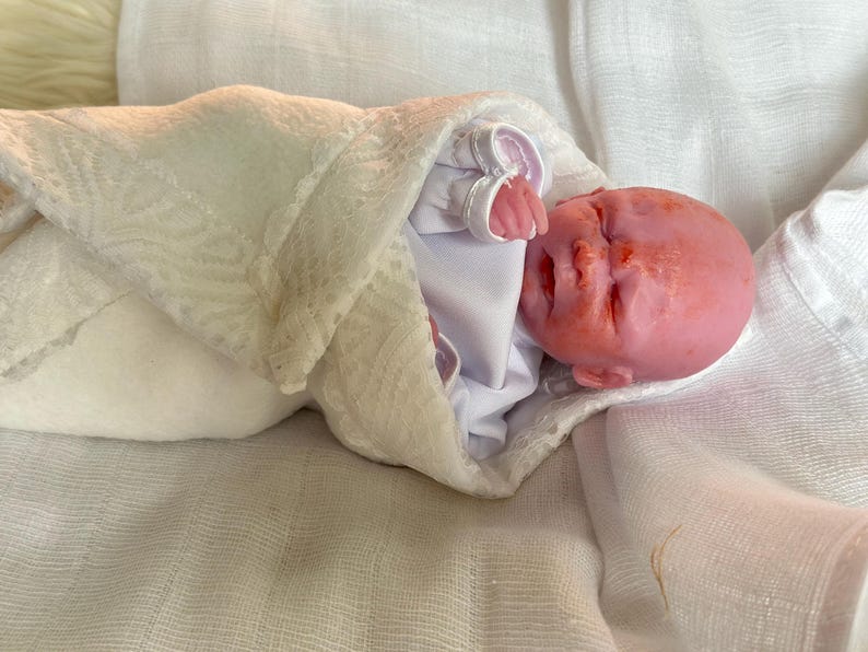 K&ouml;nnte beinhalten: Eine realistische Silikonpuppe mit rosafarbenem Teint, geschlossenen Augen und kleinem Mund, eingewickelt in ein wei&szlig;es lang&auml;rmeliges Kleidungsst&uuml;ck und eine wei&szlig;e Wickeldecke mit Spitzenbesatz, mit einem glatten, haarlosen Kopf und sichtbaren Fingern.