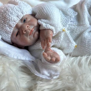May include: A realistic newborn doll with pale skin and blue eyes, wearing a white hand-knitted outfit consisting of a long-sleeved cardigan with small yellow buttons, matching trousers, and a textured beanie hat with a pom-pom on top.