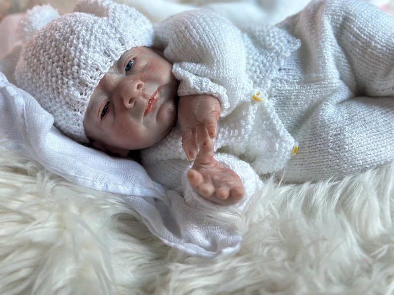 May include: A realistic newborn doll with pale skin and blue eyes, wearing a white hand-knitted outfit consisting of a long-sleeved cardigan with small yellow buttons, matching trousers, and a textured beanie hat with a pom-pom on top.