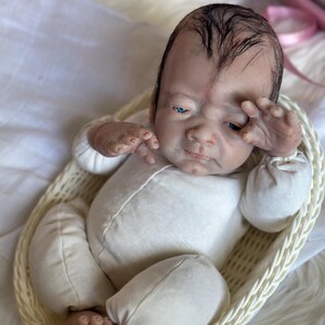 May include: A realistic reborn doll with painted dark hair, blue eyes, and a fair complexion, wearing a long-sleeved cream-colored onesie. The doll is positioned inside a light-colored woven oval basket, with one hand raised near its face.