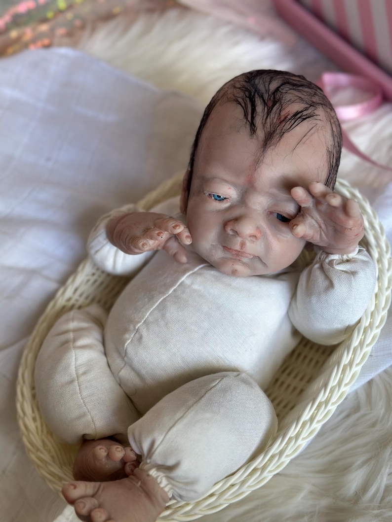 May include: A realistic reborn doll with painted dark hair, blue eyes, and a fair complexion, wearing a long-sleeved cream-colored onesie. The doll is positioned inside a light-colored woven oval basket, with one hand raised near its face.
