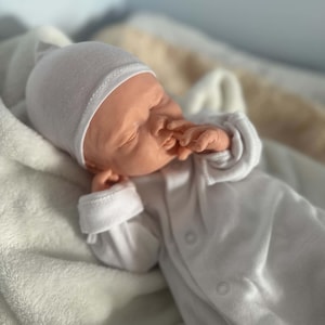 May include: A realistic newborn doll with light skin, closed eyes, and a slightly open mouth. The doll is wearing a plain white long-sleeved onesie with visible snap buttons down the front and a matching white beanie cap, resting on a soft white blanket.