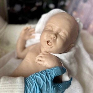 May include: A realistic baby doll with closed eyes and an open mouth, lying on a white blanket. The doll is being held by a gloved hand.