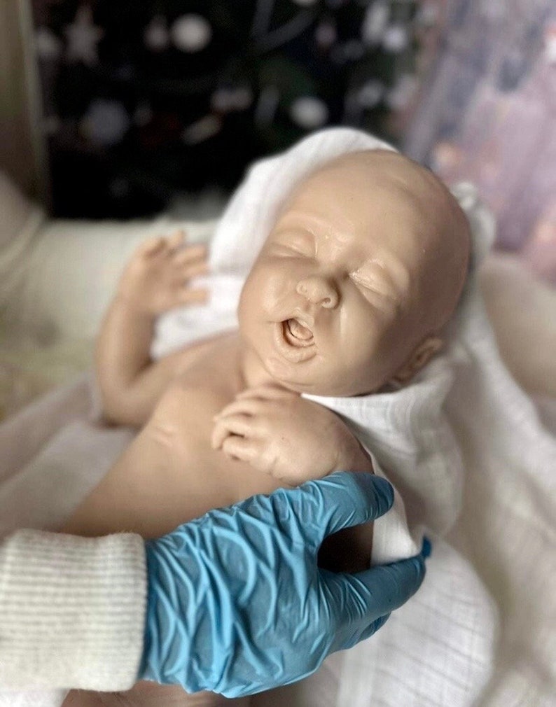 May include: A realistic baby doll with closed eyes and an open mouth, lying on a white blanket. The doll is being held by a gloved hand.