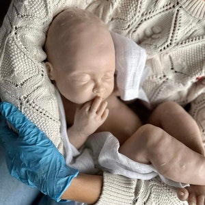 May include: A realistic baby doll with a soft, pale skin tone, wearing a white cloth diaper and sleeping peacefully in the arms of a person wearing a cream-colored knitted sweater.