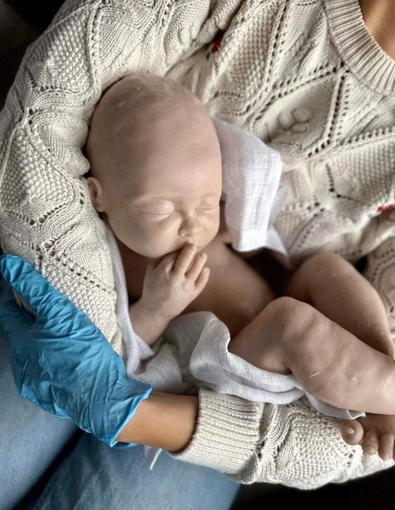 May include: A realistic baby doll with a soft, pale skin tone, wearing a white cloth diaper and sleeping peacefully in the arms of a person wearing a cream-colored knitted sweater.