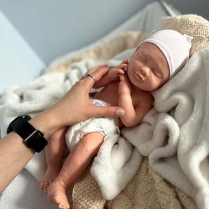 May include: A realistic newborn baby doll with light skin, closed eyes, and a soft expression. The doll wears a plain white knit cap and a disposable diaper with colorful patterns. It is resting on a textured cream-colored blanket with a blue pacifier nearby.