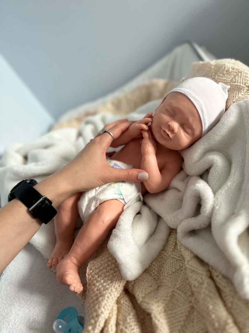 May include: A realistic newborn baby doll with light skin, closed eyes, and a soft expression. The doll wears a plain white knit cap and a disposable diaper with colorful patterns. It is resting on a textured cream-colored blanket with a blue pacifier nearby.