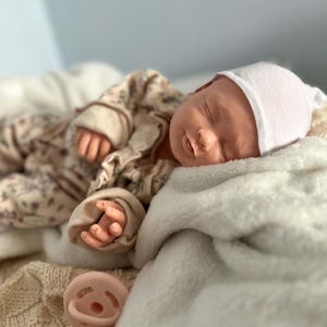 May include: A realistic newborn doll with closed eyes, wearing a white beanie and a beige long-sleeved outfit with a subtle floral pattern. The doll is resting on a soft white blanket, with a light pink pacifier placed nearby on a textured fabric.