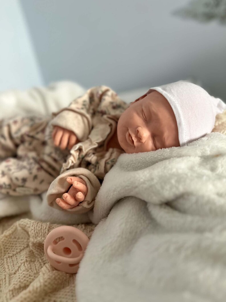 May include: A realistic newborn doll with closed eyes, wearing a white beanie and a beige long-sleeved outfit with a subtle floral pattern. The doll is resting on a soft white blanket, with a light pink pacifier placed nearby on a textured fabric.