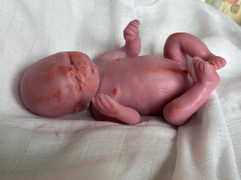 K&ouml;nnte beinhalten: Eine Miniatur-Silikonpuppe, die einem Neugeborenen nachempfunden ist, mit rosafarbenen Hautt&ouml;nen und subtilen r&ouml;tlichen Flecken im Gesicht, auf der Brust und an den Gliedma&szlig;en. Die Figur ist in einer liegenden Position mit angewinkelten Armen und Beinen.