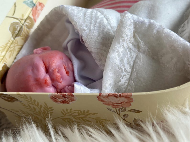K&ouml;nnte beinhalten: Eine kleine, rosafarbene Puppe mit geschlossenen Augen und texturiertem Gesicht, eingewickelt in eine wei&szlig;e Stoffdecke mit Spitzenbesatz. Die Puppe liegt in einer rechteckigen Schachtel mit cremefarbenem Hintergrund und einem Muster aus rosa Rosen und gr&uuml;nen Bl&auml;ttern.