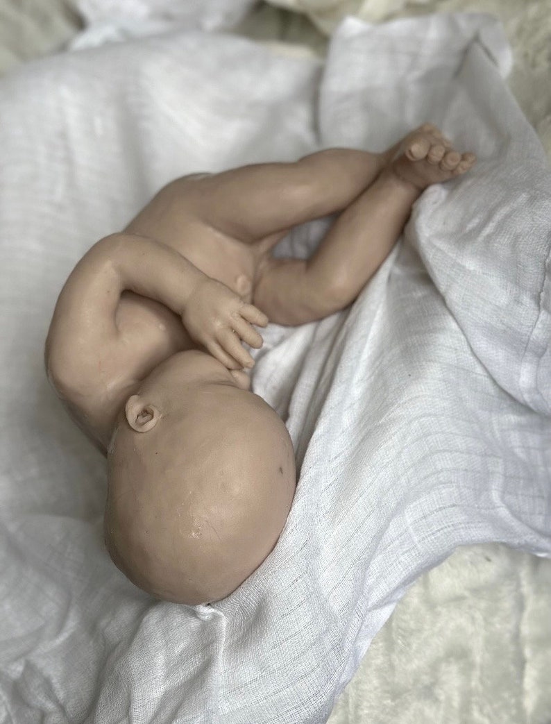 K&ouml;nnte beinhalten: Eine realistische, nackte Babypuppe liegt auf einem wei&szlig;en, strukturierten Stoff. Die Puppe befindet sich in einer F&ouml;tusstellung, mit zur Seite geneigtem Kopf und leicht angewinkelten Armen und Beinen. Der Hautton ist hellbeige.