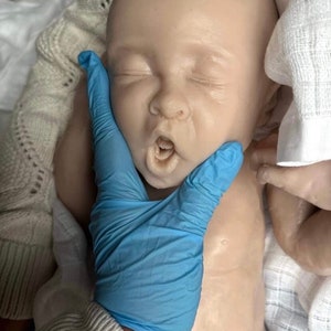 May include: A realistic baby doll with closed eyes and an open mouth, wearing a white fabric. A gloved hand is gently touching the doll's cheek.