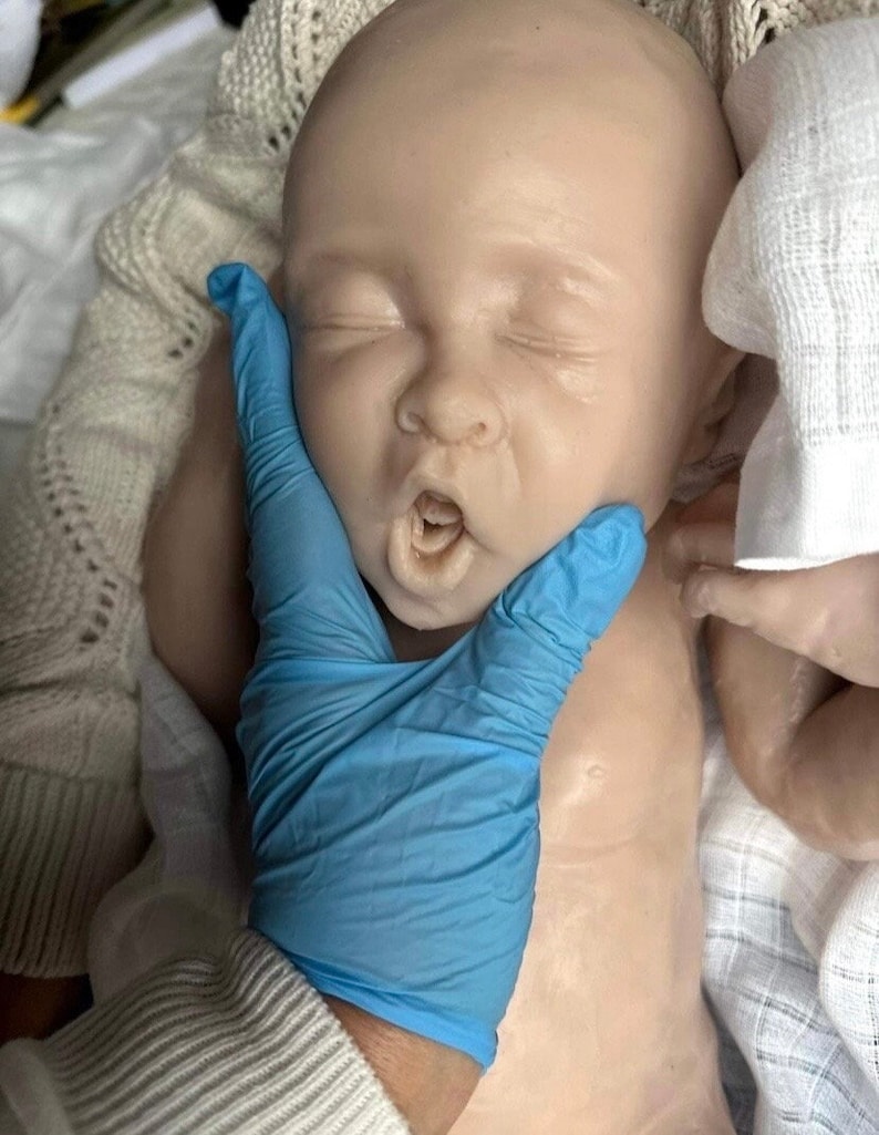 May include: A realistic baby doll with closed eyes and an open mouth, wearing a white fabric. A gloved hand is gently touching the doll's cheek.