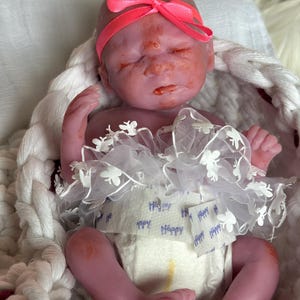K&ouml;nnte beinhalten: Eine Miniatur-Silikonpuppe mit rosigem Hautton, die mit geschlossenen Augen schl&auml;ft. Die Puppe tr&auml;gt ein leuchtend neonpinkes Stirnband, eine wei&szlig;e Windel mit dem wiederholt aufgedruckten Wort Happy und einen wei&szlig;en T&uuml;llrock mit kleinen Blumen.