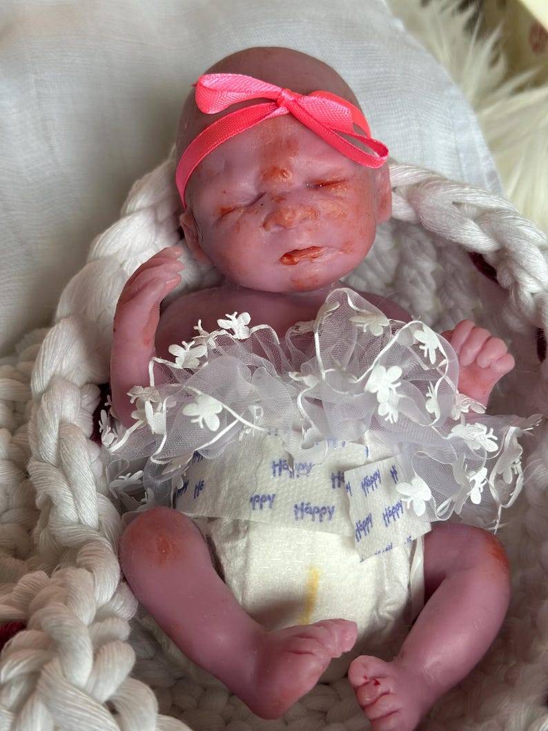 K&ouml;nnte beinhalten: Eine Miniatur-Silikonpuppe mit rosigem Hautton, die mit geschlossenen Augen schl&auml;ft. Die Puppe tr&auml;gt ein leuchtend neonpinkes Stirnband, eine wei&szlig;e Windel mit dem wiederholt aufgedruckten Wort Happy und einen wei&szlig;en T&uuml;llrock mit kleinen Blumen.