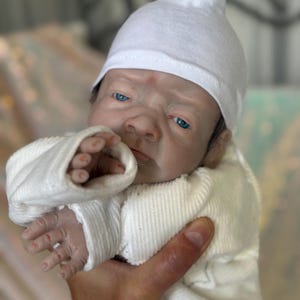 May include: A realistic reborn doll with pale skin, bright blue eyes, and detailed facial features. The doll wears a plain white long-sleeved ribbed shirt and a matching white knit cap. The doll has visible fingers and a lifelike expression.