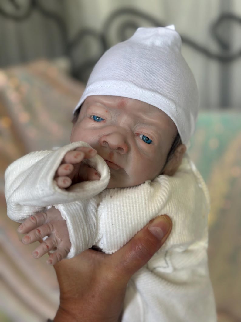May include: A realistic reborn doll with pale skin, bright blue eyes, and detailed facial features. The doll wears a plain white long-sleeved ribbed shirt and a matching white knit cap. The doll has visible fingers and a lifelike expression.