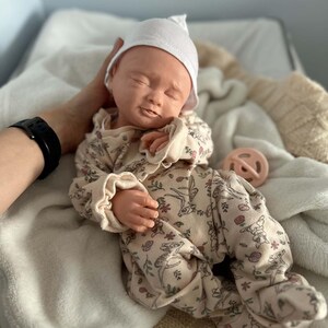 May include: A realistic baby doll with closed eyes and a neutral expression, wearing a white knit cap and a beige long-sleeved footed sleeper featuring a repeating pattern of small deer and floral motifs, accompanied by a round light pink pacifier.