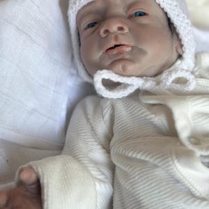 May include: A realistic baby doll with pale skin, blue eyes, and dark hair, wearing a white ribbed long-sleeve onesie and a white crocheted bonnet with a tie closure. The doll has detailed facial features and is positioned on a soft white fabric.