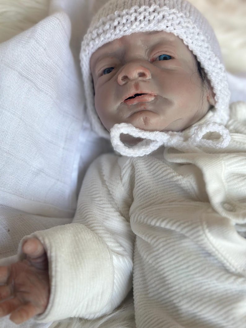 May include: A realistic baby doll with pale skin, blue eyes, and dark hair, wearing a white ribbed long-sleeve onesie and a white crocheted bonnet with a tie closure. The doll has detailed facial features and is positioned on a soft white fabric.