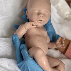 Puede incluir: Un mu&ntilde;eco de beb&eacute; realista de silicona con los ojos cerrados, la boca ligeramente entreabierta y un tono de piel marr&oacute;n claro. El mu&ntilde;eco est&aacute; siendo sostenido por manos enguantadas.