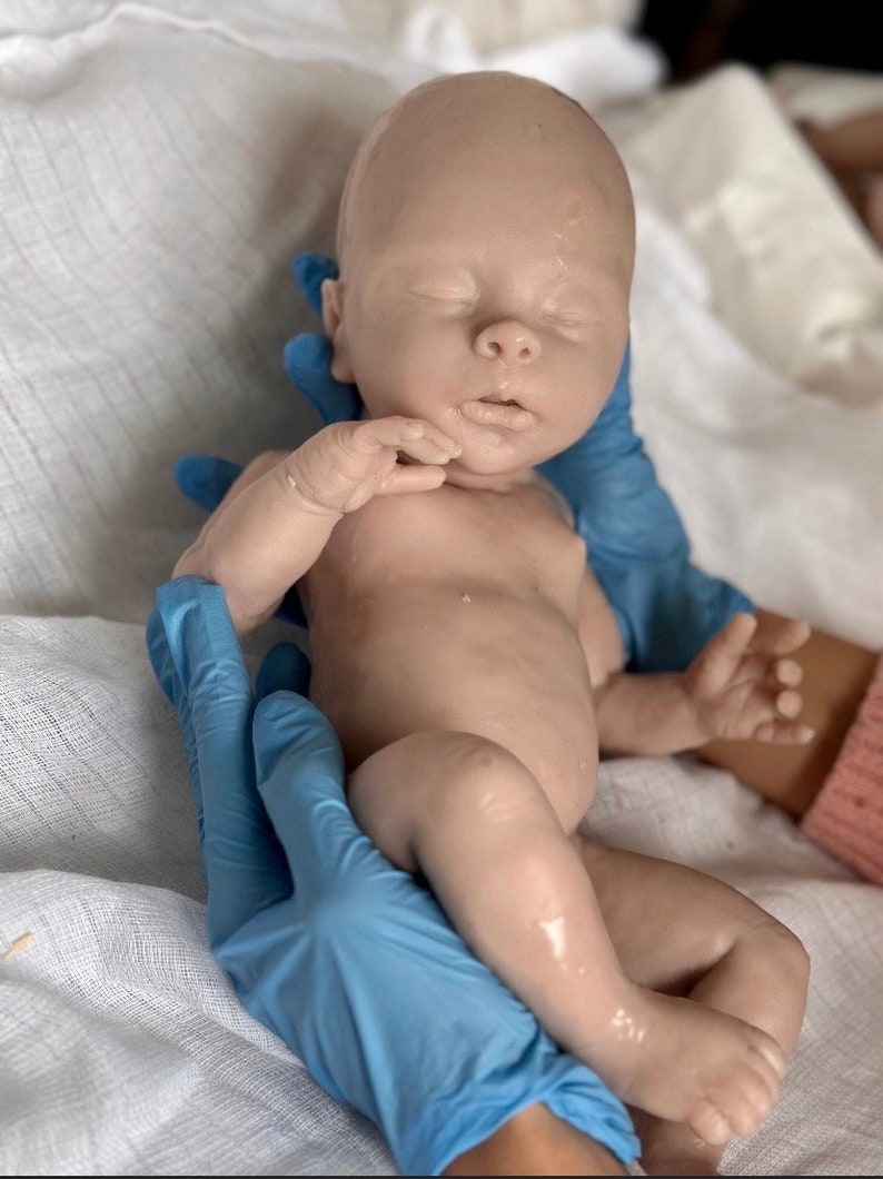 Puede incluir: Un mu&ntilde;eco de beb&eacute; realista de silicona con los ojos cerrados, la boca ligeramente entreabierta y un tono de piel marr&oacute;n claro. El mu&ntilde;eco est&aacute; siendo sostenido por manos enguantadas.