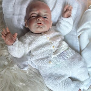 May include: A realistic reborn doll with a bald head and detailed skin texture, wearing a white knitted long-sleeve cardigan with small buttons and matching white knitted pants. The doll is positioned with arms raised, resting on a soft white fabric.