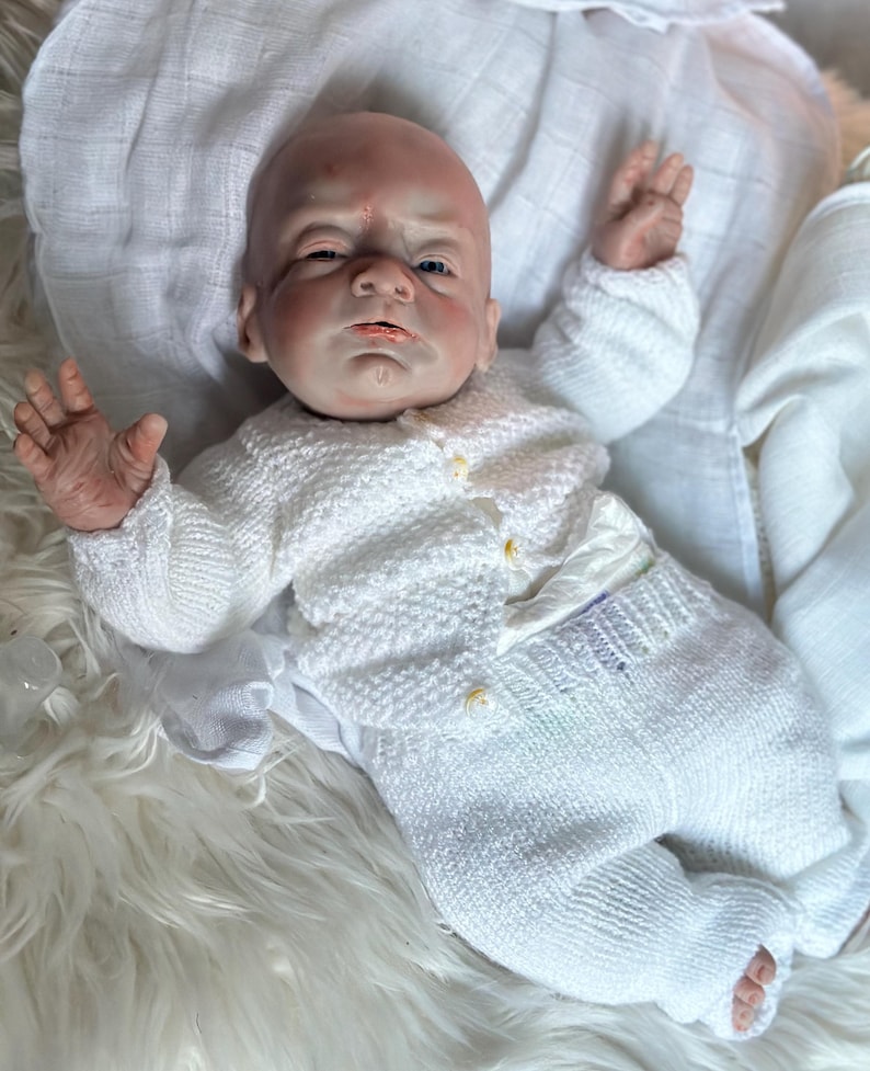 May include: A realistic reborn doll with a bald head and detailed skin texture, wearing a white knitted long-sleeve cardigan with small buttons and matching white knitted pants. The doll is positioned with arms raised, resting on a soft white fabric.