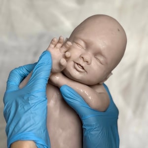 K&ouml;nnte beinhalten: Eine lebensechte Babypuppe mit heller Haut, geschlossenen Augen und einem leichten L&auml;cheln. Der Arm der Puppe ist erhoben, und ihre Hand ist in der N&auml;he ihres Gesichts. Die Puppe wird von zwei H&auml;nden mit blauen Handschuhen gehalten.