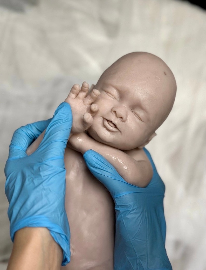 K&ouml;nnte beinhalten: Eine lebensechte Babypuppe mit heller Haut, geschlossenen Augen und einem leichten L&auml;cheln. Der Arm der Puppe ist erhoben, und ihre Hand ist in der N&auml;he ihres Gesichts. Die Puppe wird von zwei H&auml;nden mit blauen Handschuhen gehalten.