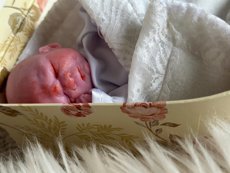 K&ouml;nnte beinhalten: Eine kleine, rosafarbene Puppe mit geschlossenen Augen und detaillierten Gesichtsz&uuml;gen, die in einer cremefarbenen Schachtel mit Rosenmuster liegt. Die Puppe ist teilweise mit einem wei&szlig;en, halbtransparenten Stoff mit zarter Spitzenstruktur bedeckt.