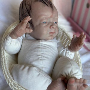 May include: A realistic reborn doll with pale skin, blue eyes, and dark painted hair, wearing a long-sleeved white onesie. The doll is positioned inside a small, oval-shaped white woven basket, with its arms raised and legs slightly bent.