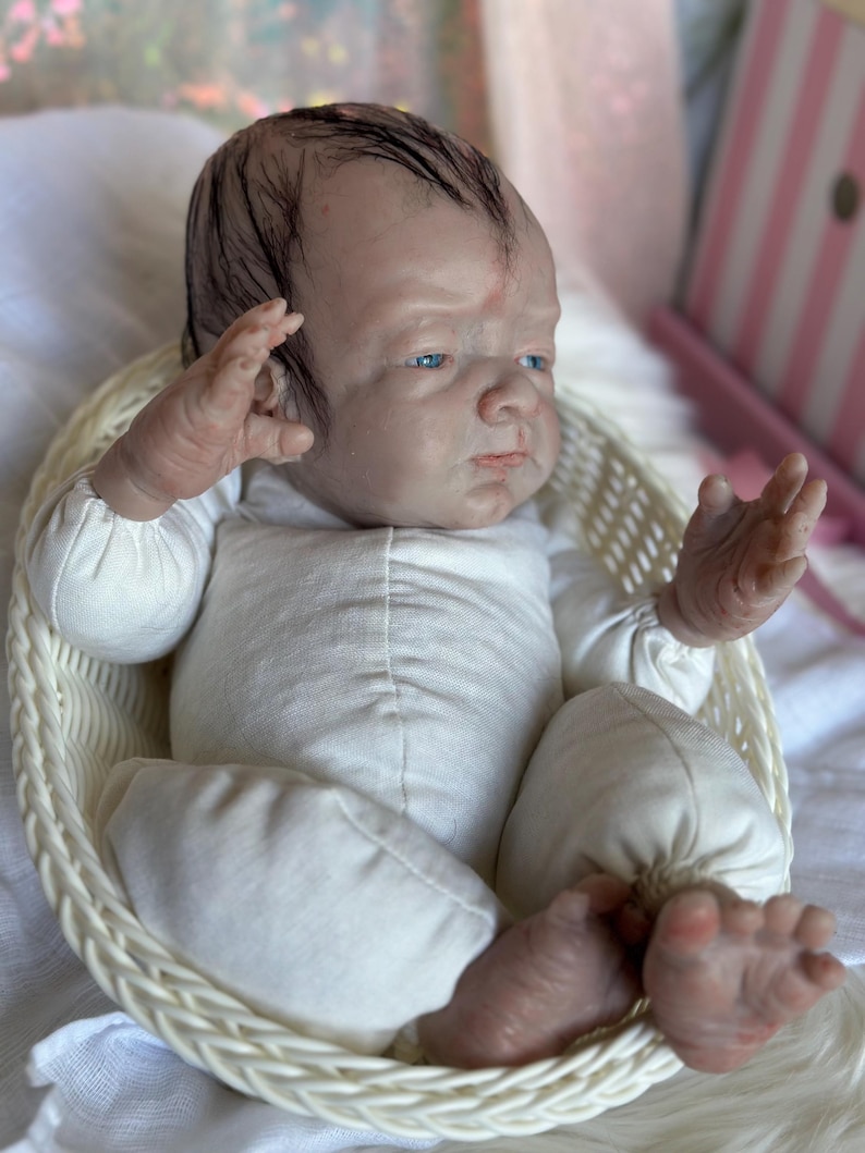 May include: A realistic reborn doll with pale skin, blue eyes, and dark painted hair, wearing a long-sleeved white onesie. The doll is positioned inside a small, oval-shaped white woven basket, with its arms raised and legs slightly bent.