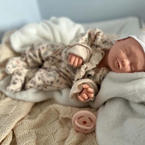 May include: A realistic newborn doll with closed eyes, wearing a white cap and a beige long-sleeved onesie featuring a subtle floral pattern. The doll is resting on a soft, textured cream-colored blanket, accompanied by a small, round, pale pink pacifier.