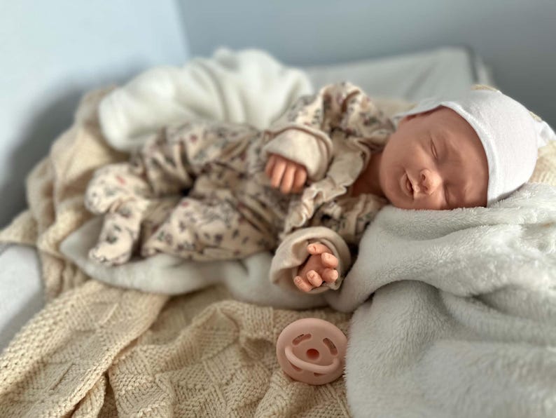 May include: A realistic newborn doll with closed eyes, wearing a white cap and a beige long-sleeved onesie featuring a subtle floral pattern. The doll is resting on a soft, textured cream-colored blanket, accompanied by a small, round, pale pink pacifier.