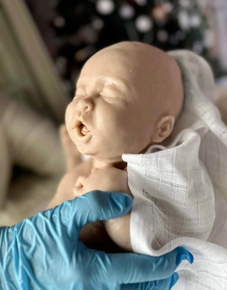 May include: A realistic baby doll with closed eyes and an open mouth, made of soft silicone. The doll is wrapped in a white cloth and being held by a gloved hand.