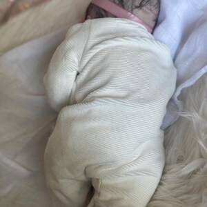 May include: A realistic baby doll with dark hair and a pink headband, wearing a long-sleeved, footed, cream-colored ribbed onesie. The doll is positioned lying on its stomach, showcasing the textured fabric of the garment and the detailed hair.