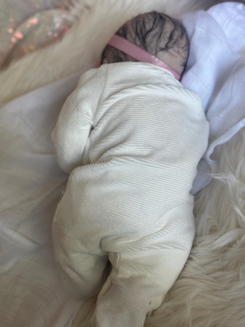 May include: A realistic baby doll with dark hair and a pink headband, wearing a long-sleeved, footed, cream-colored ribbed onesie. The doll is positioned lying on its stomach, showcasing the textured fabric of the garment and the detailed hair.