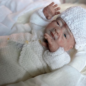 May include: A realistic newborn doll with light skin and blue eyes, wearing a white knitted cardigan, matching white knitted pants, and a white textured knitted hat with a small pom-pom on top. The doll is lying down with arms raised.