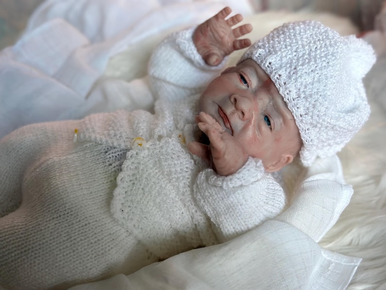 May include: A realistic newborn doll with light skin and blue eyes, wearing a white knitted cardigan, matching white knitted pants, and a white textured knitted hat with a small pom-pom on top. The doll is lying down with arms raised.