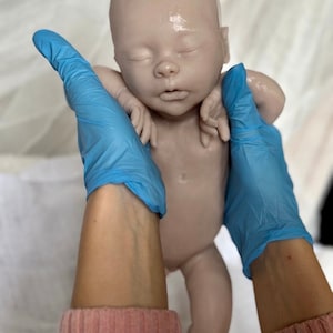 Puede incluir: Un mu&ntilde;eco de beb&eacute; de silicona realista con una tez rosa claro, ojos cerrados y boca ligeramente abierta. El mu&ntilde;eco est&aacute; siendo sostenido por manos enguantadas.